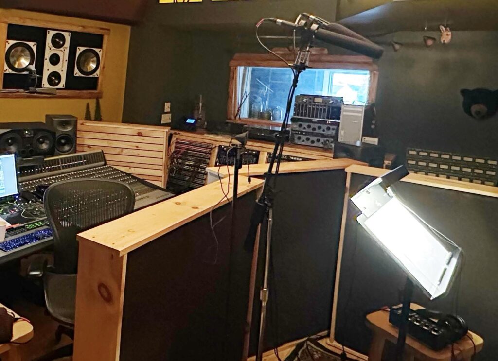 A cozy recording studio features a mixing console with speakers, a microphone on a stand, and a music stand with light. The room feels warm and creative.