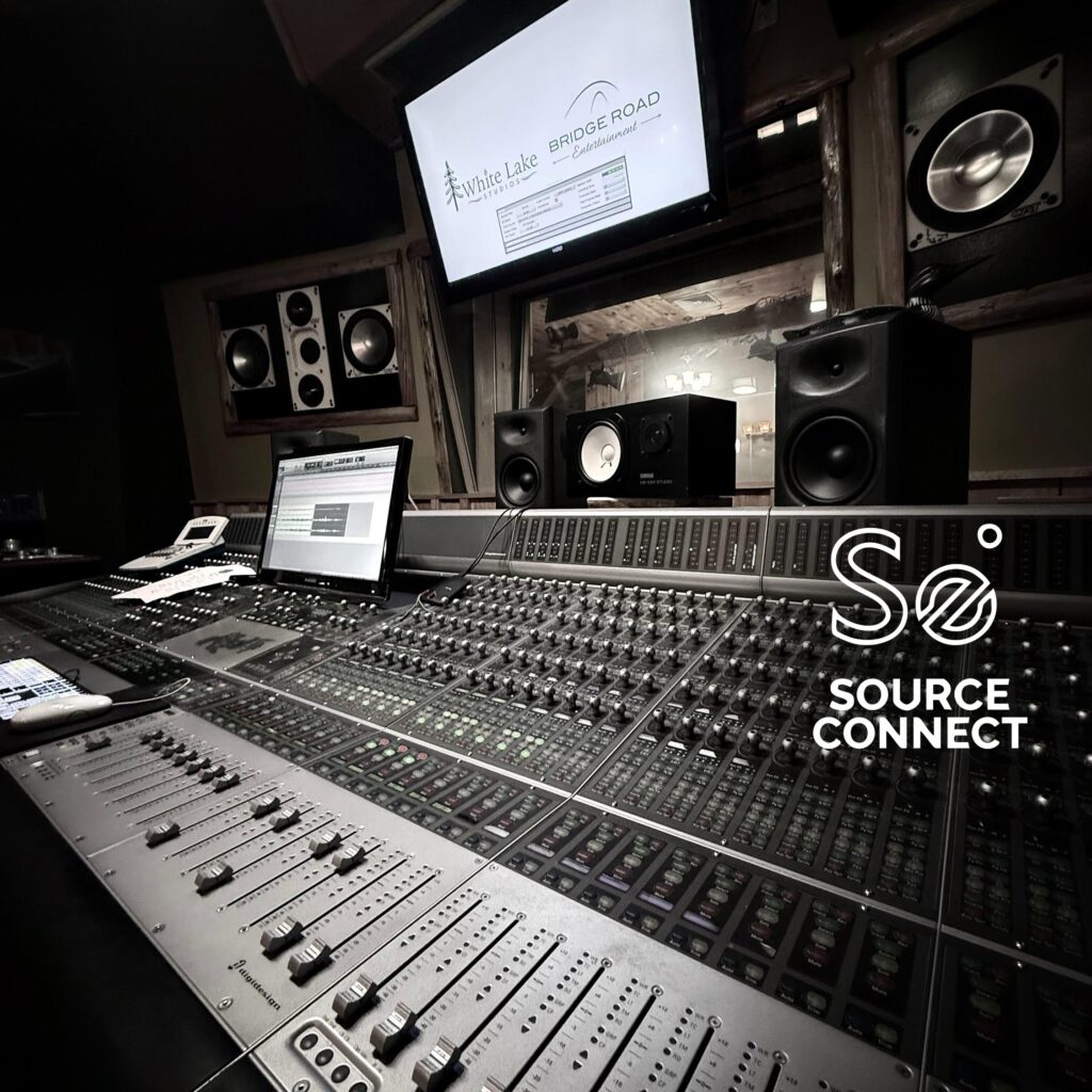 A professional audio mixing console with multiple faders and control panels, speakers, monitors, and a screen displaying "White Lake Bridge Road" in a recording studio.
