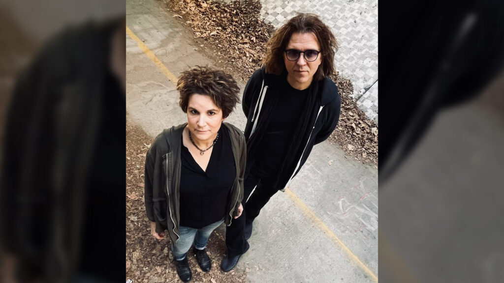 Little Days vocalist Mini Diaz and bassist Jorgen Carlsson back stand on a leaf-strewn path, looking up. Both wear dark clothing and serious expressions. The background includes scattered autumn leaves.