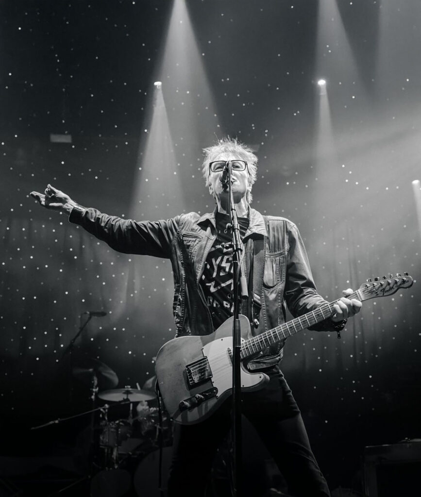 Black and white image of guitarist Tommy Stinson singing passionately on stage, arm raised. Spotlight beams and a drum set are visible, creating an energetic, lively scene.